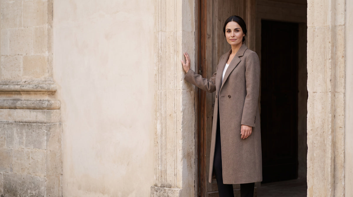 
Sofia wears a taupe coat beside a wooden door, in the light of a stone courtyard.